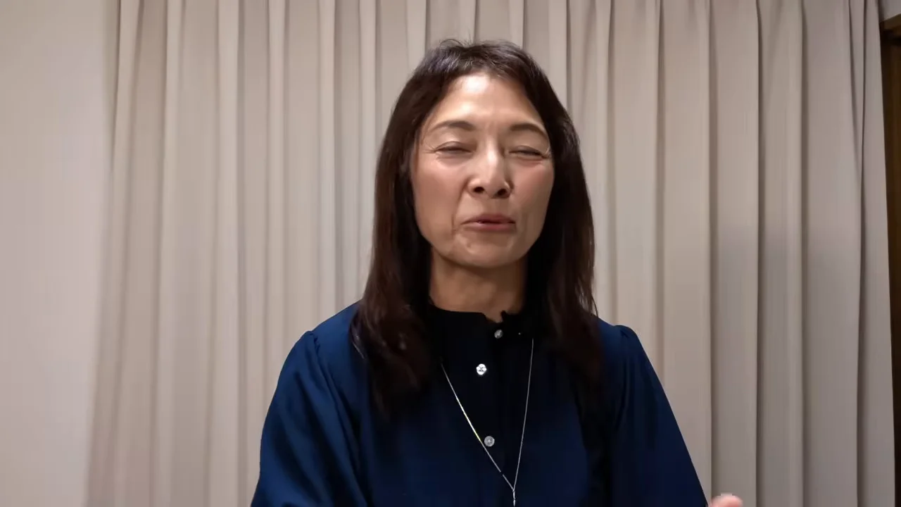 カーテンの前に立つ女性がカメラに向かって話しています。