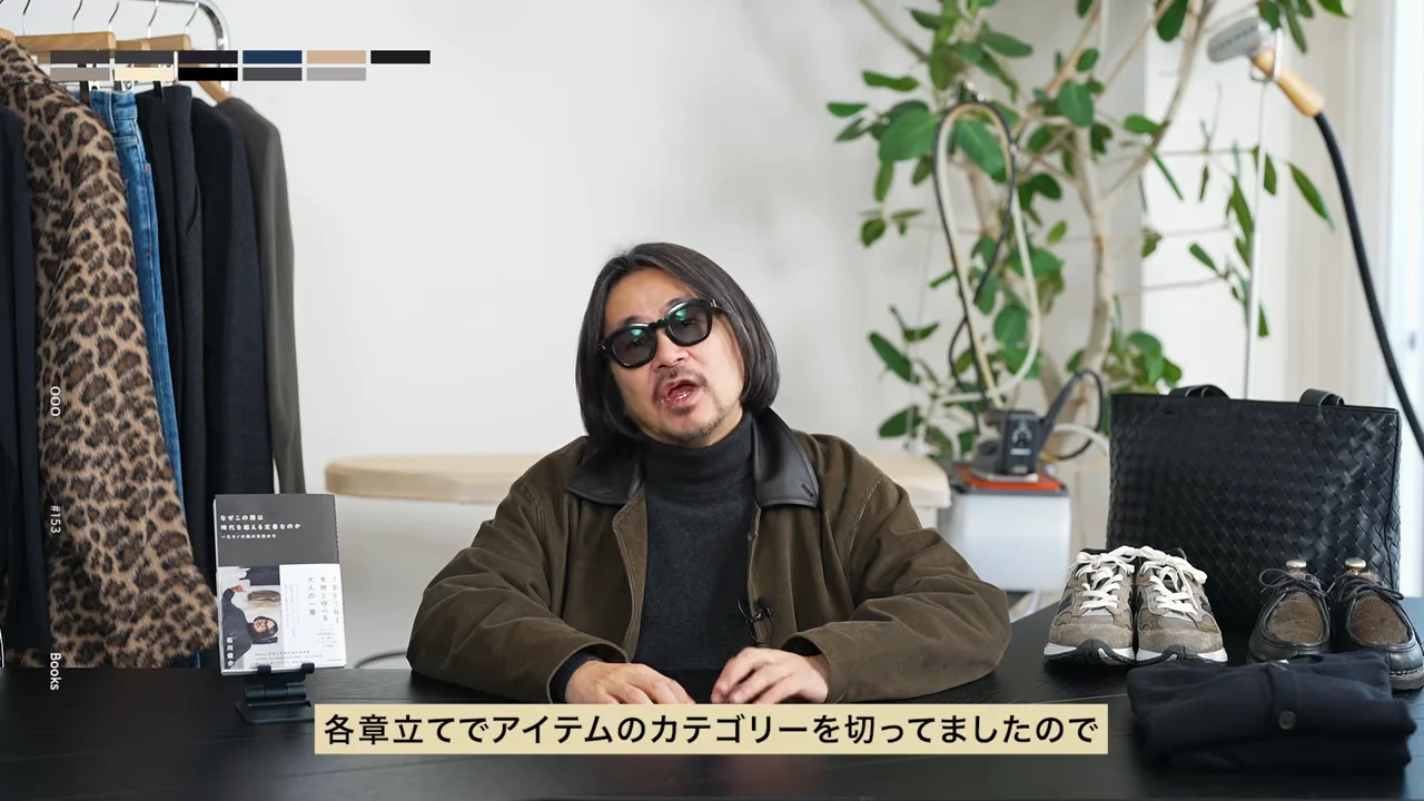 サングラスをかけた男性が机の前に座り、本や靴、バッグなどの愛用品が並べられています。