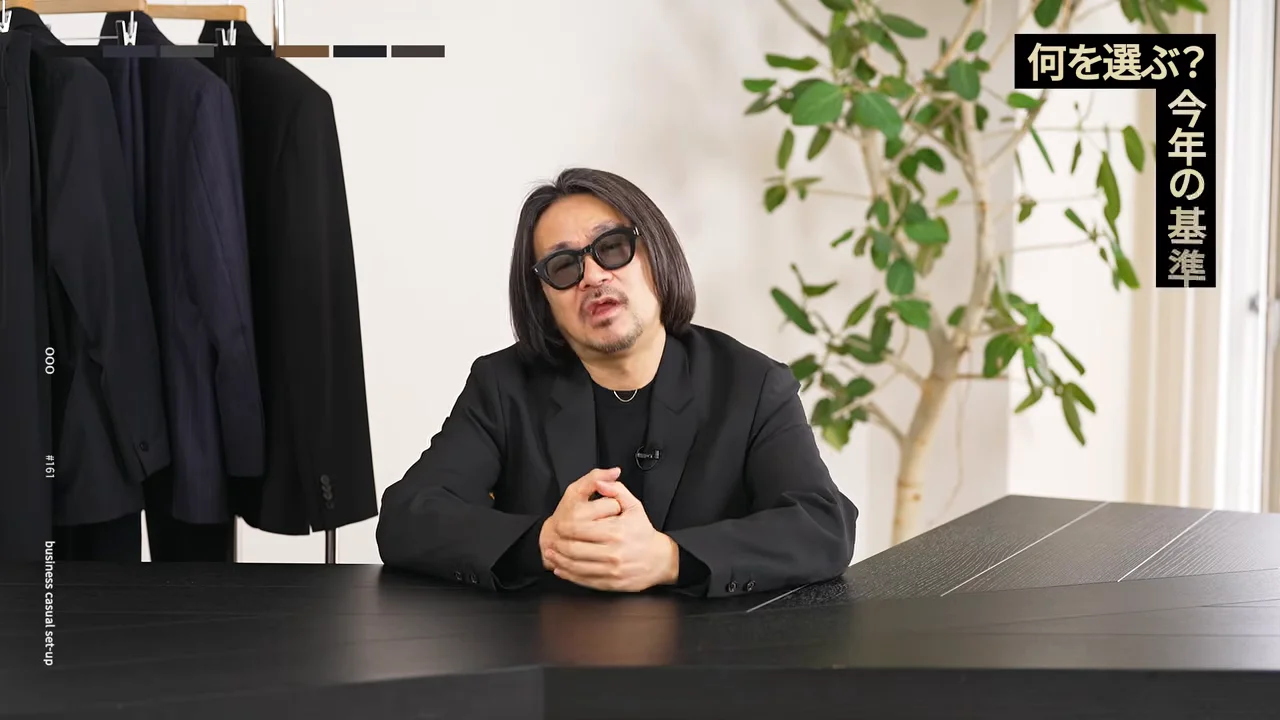 黒いジャケットを着てサングラスをかけた男性が、テーブルの前に座って話しています。