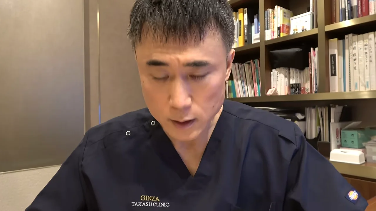 本棚を背にして紺色のスクラブを着た男性が、視線を落として座っている姿。
