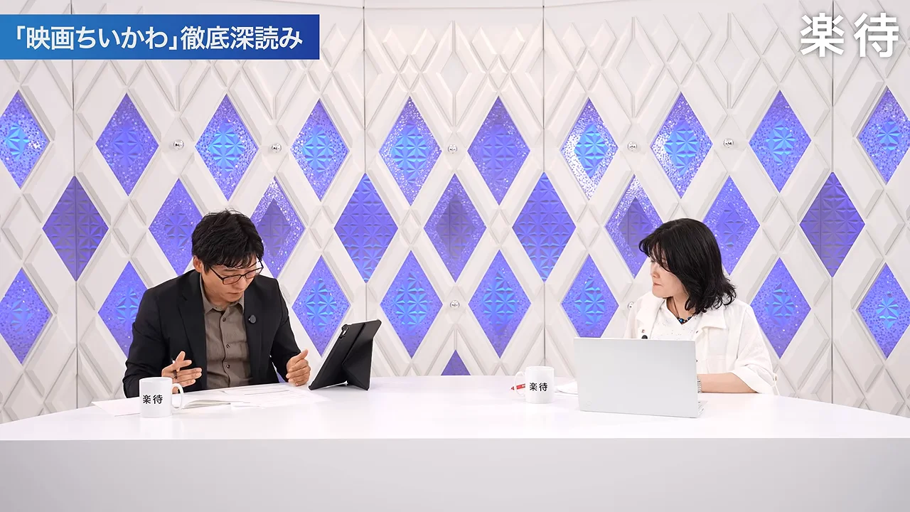 スタジオの白いデスクに向かい合って座る男性と女性の引きのショット。