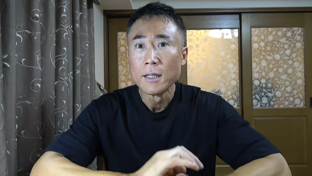 黒いTシャツを着た男性が、カーテンと引き戸のある部屋でカメラに向かって話している様子。