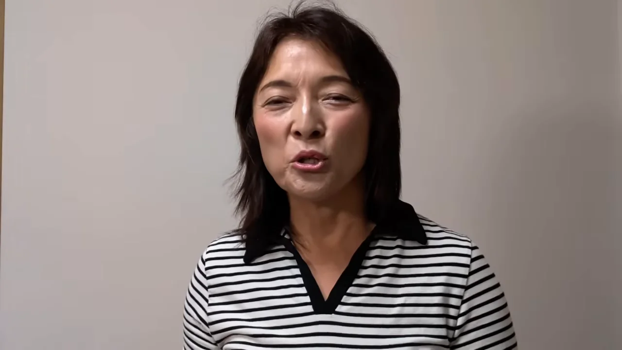 白い壁を背景に、ボーダー柄のシャツを着た女性がカメラに向かって話しています。