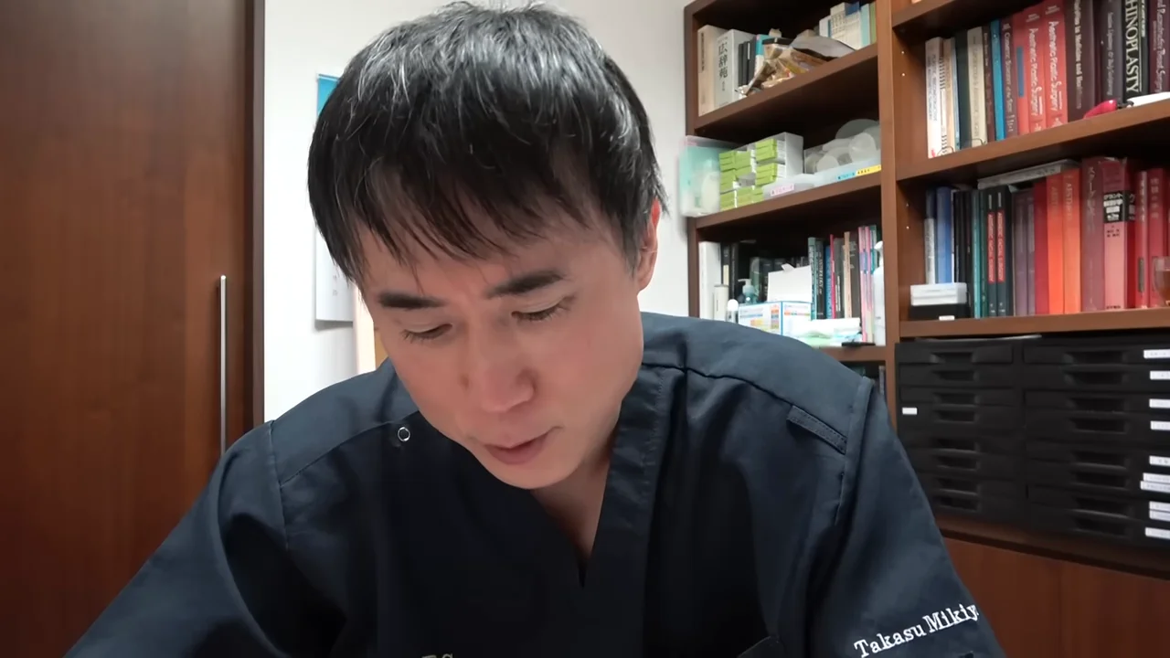 本棚のある部屋で、黒いスクラブを着た男性がうつむき加減に視線を落としています。