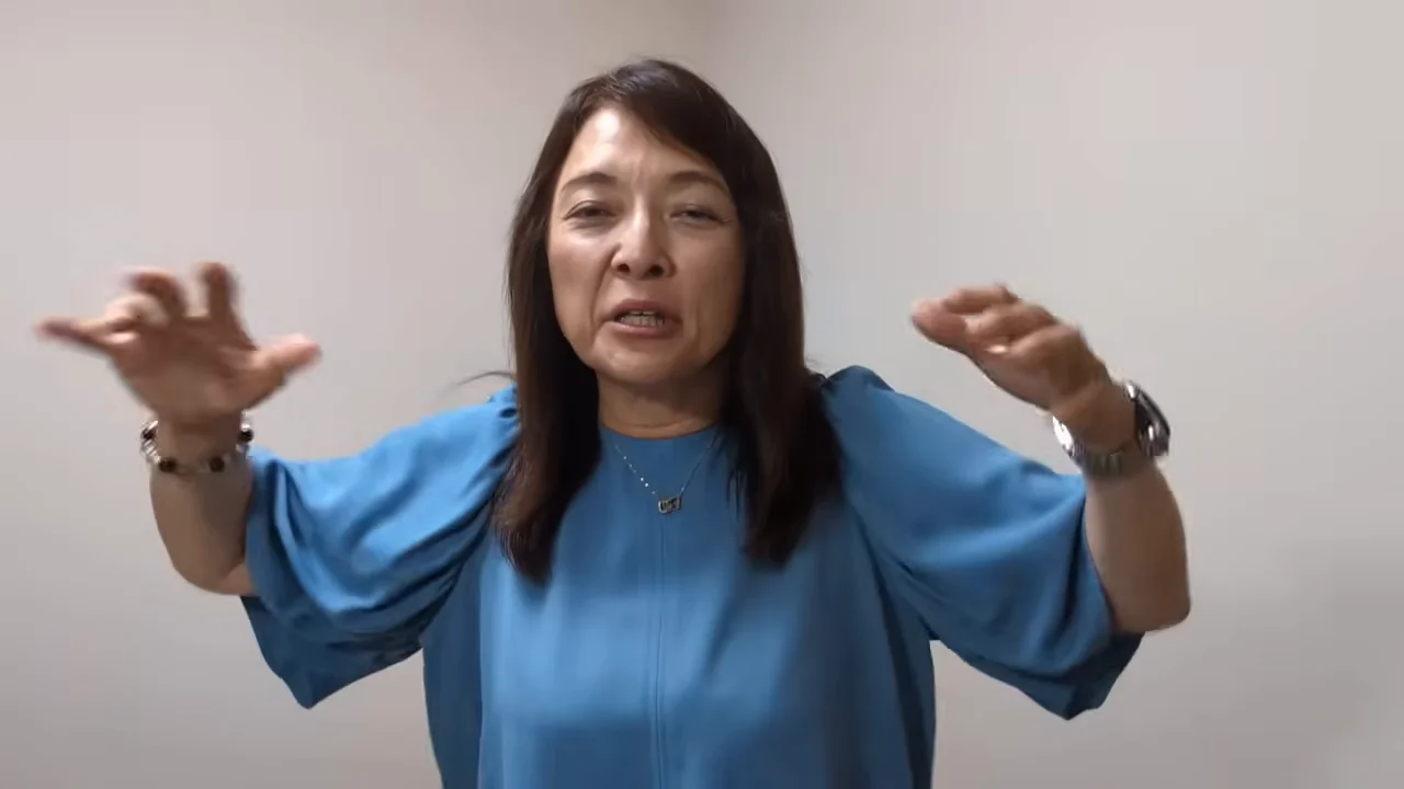 青い服を着た女性が両手を広げてジェスチャーを交えながら話している。