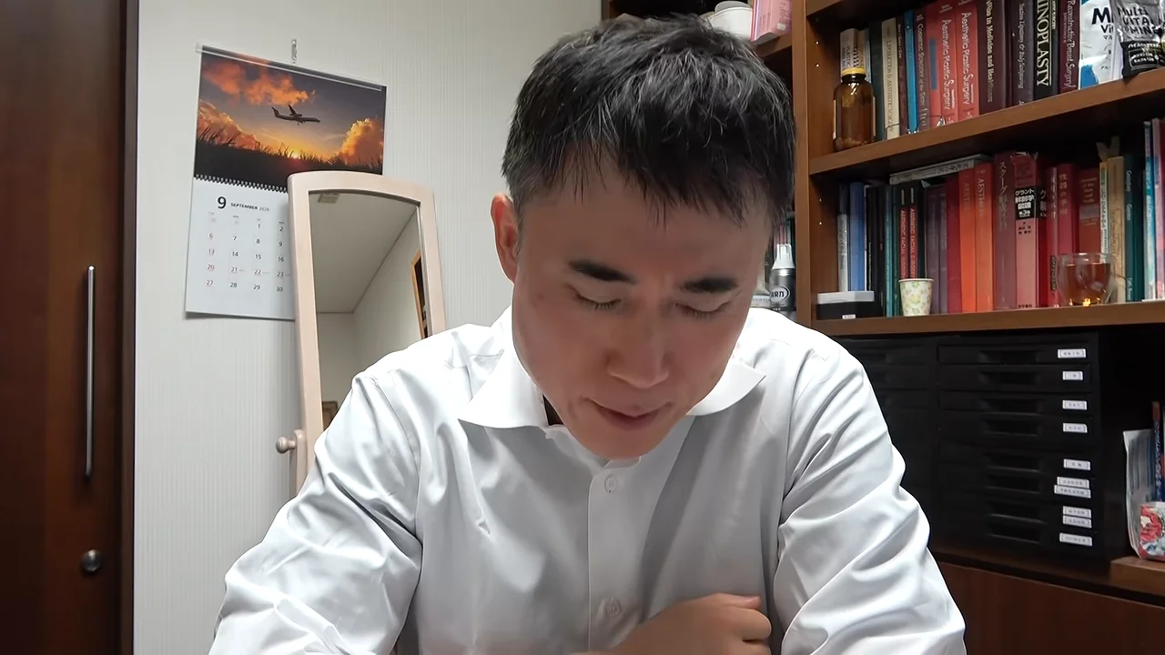 白いワイシャツを着た男性が本棚のある室内で下を向いている様子。
