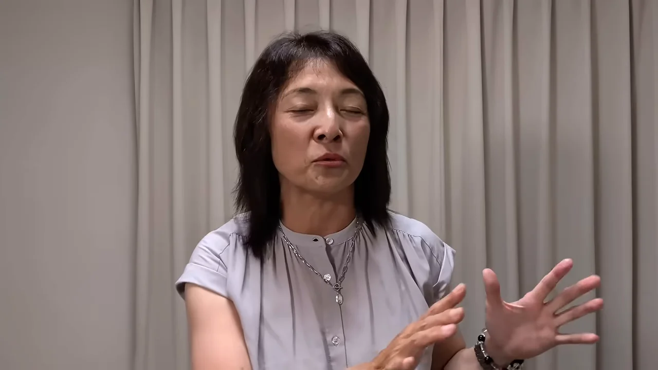 カーテンを背にした女性が、目を閉じて手ぶりを交えながら話しています。