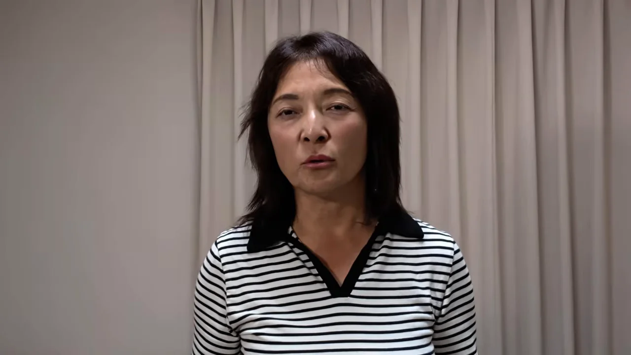 カーテンの前に立つボーダー柄の服を着た女性がカメラに向かって話しています。