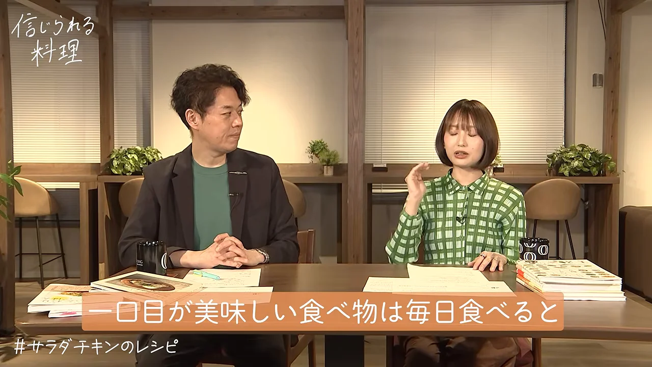 スタジオの机に向かい合い、緑色のチェック柄シャツを着た女性が手を動かしながら話している様子