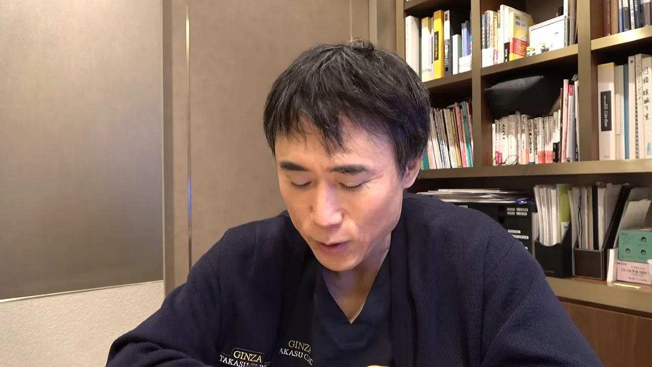本棚を背景に、うつむきながら話している男性。