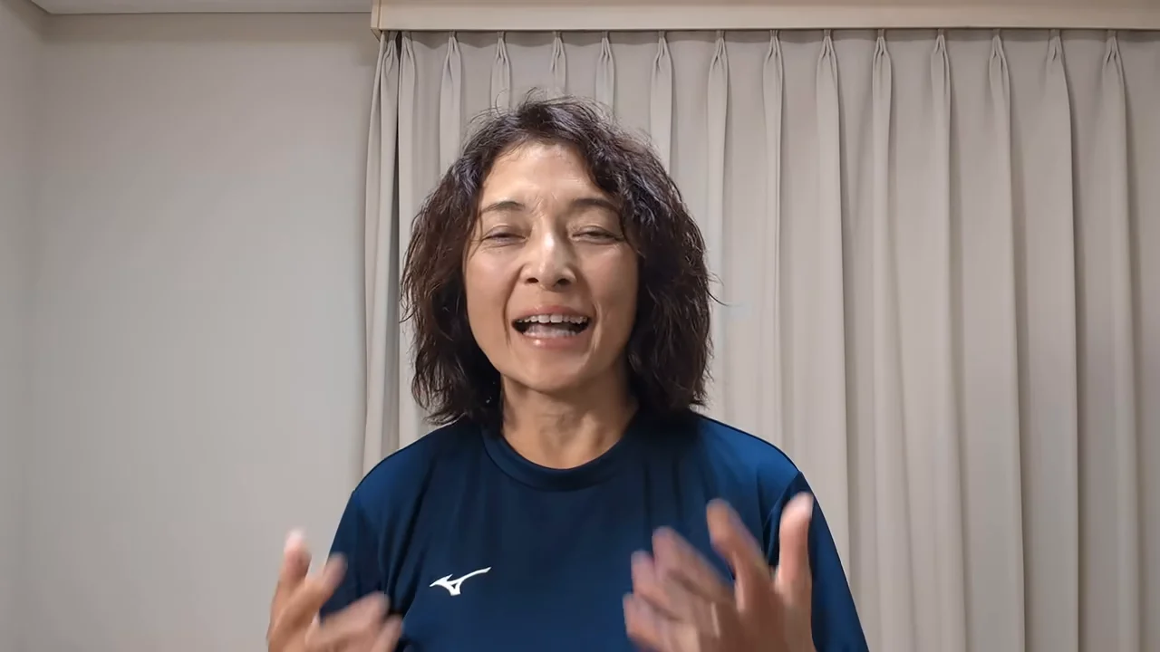 青いシャツを着た女性が、室内のカーテンの前で笑顔で身振り手振りを交えながら話している様子。