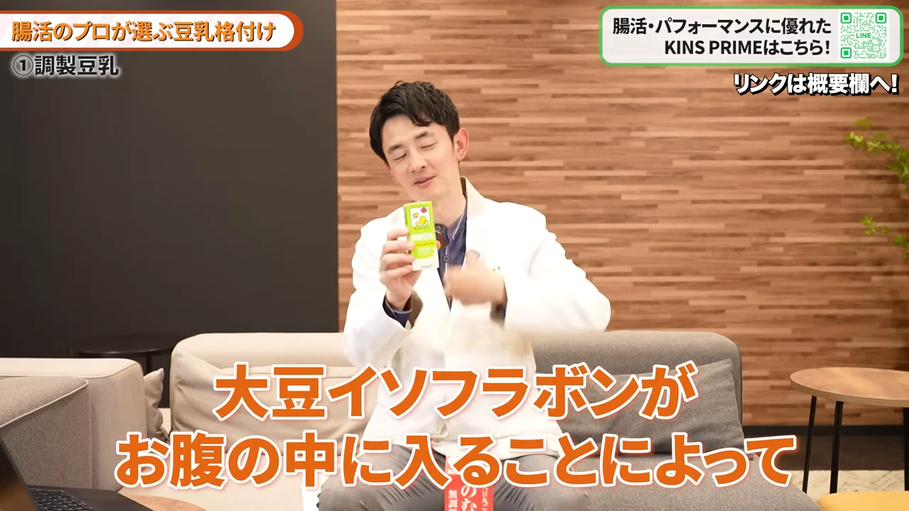 白い白衣を着た男性がソファに座り、小さな紙パックを手に持ちながら身振り手振りを交えて話している。