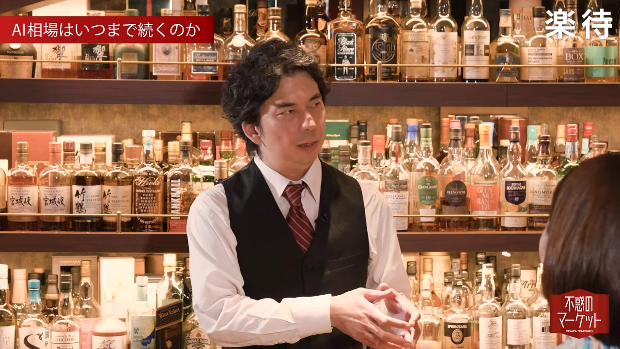 酒瓶が並ぶ棚の前で身振り手振りを交えて話すバーテンダー姿の男性