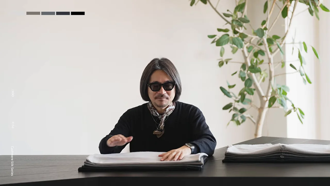 サングラスをかけた人が、テーブルの上の白い衣類に手を添え、右側には衣類が重ねて置かれている。