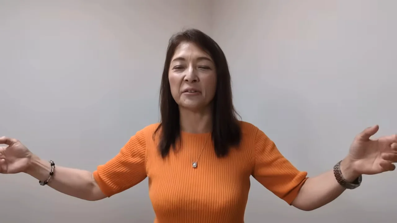 オレンジ色のトップスを着た女性が両手を広げて話している姿。