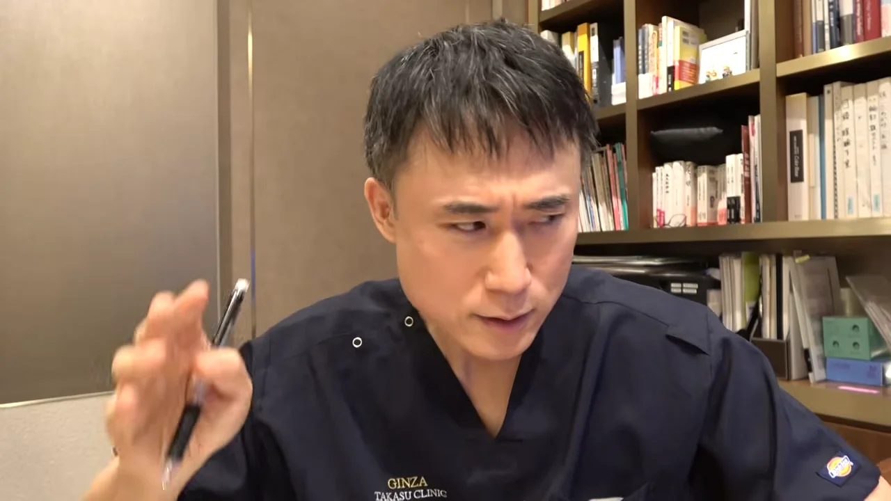 本棚の前で、濃紺の服を着た男性がペンを持った手を上げている。