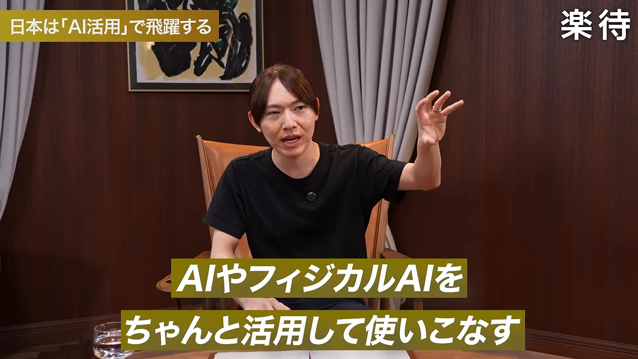 椅子に座った人物が片手を挙げながら話している様子。