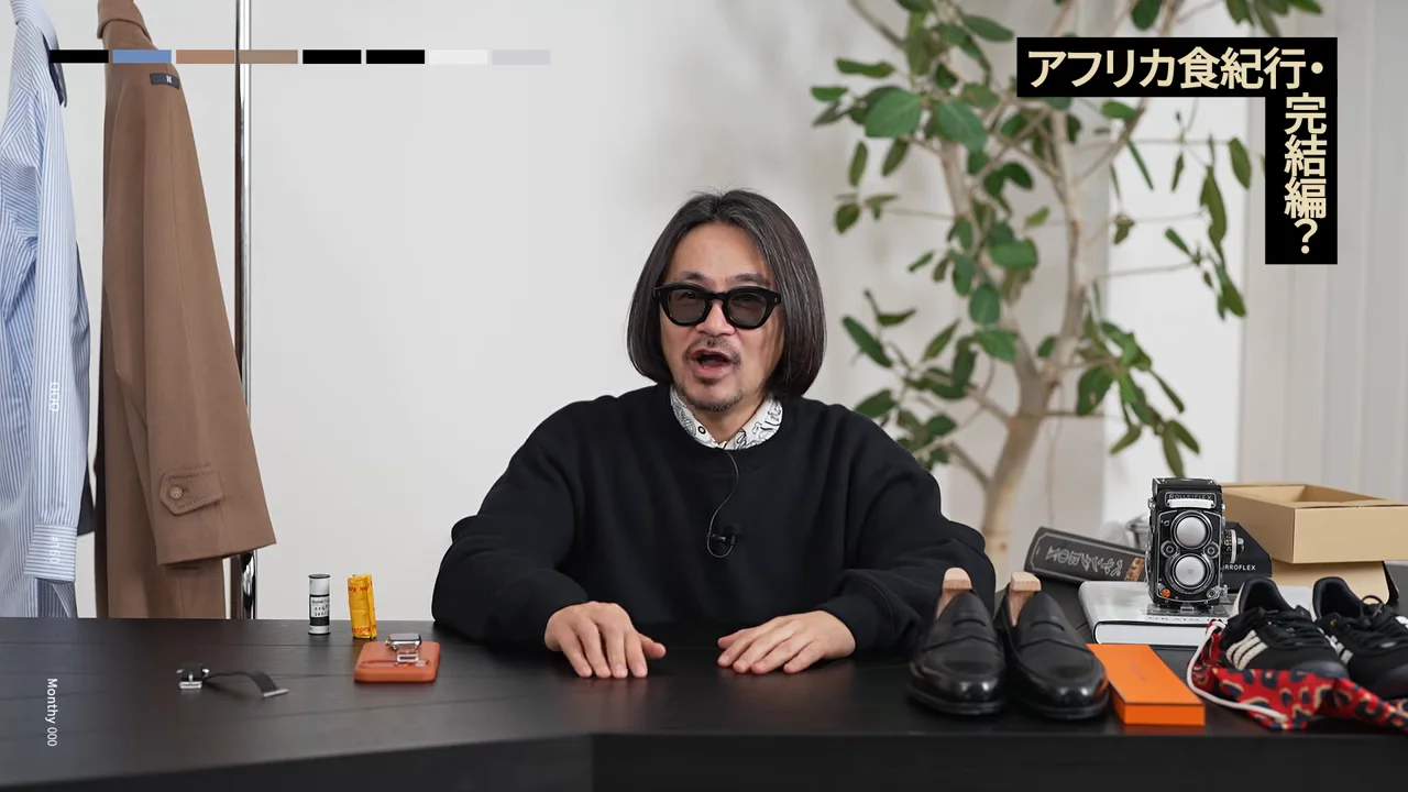 靴やカメラなどの小物が並ぶ机の前に座って話す男性