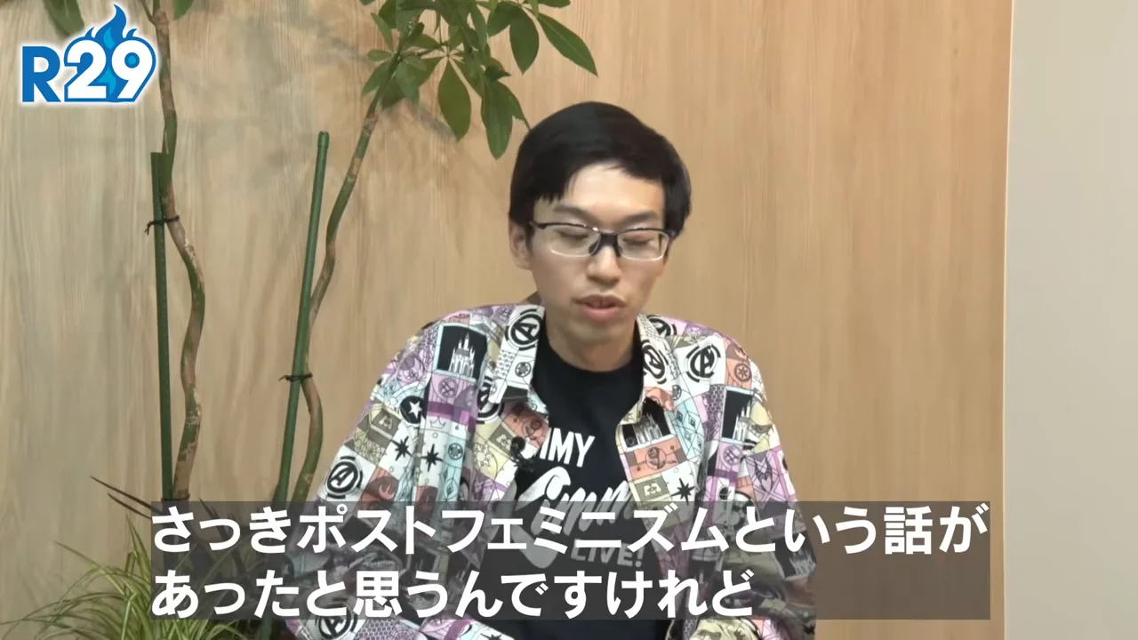 木目調の背景の前で、眼鏡をかけ柄シャツを着た男性が話している様子