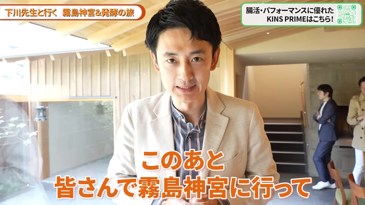 ベージュのジャケットを着た男性が、木目調の室内でカメラに向かって正面から話している様子。