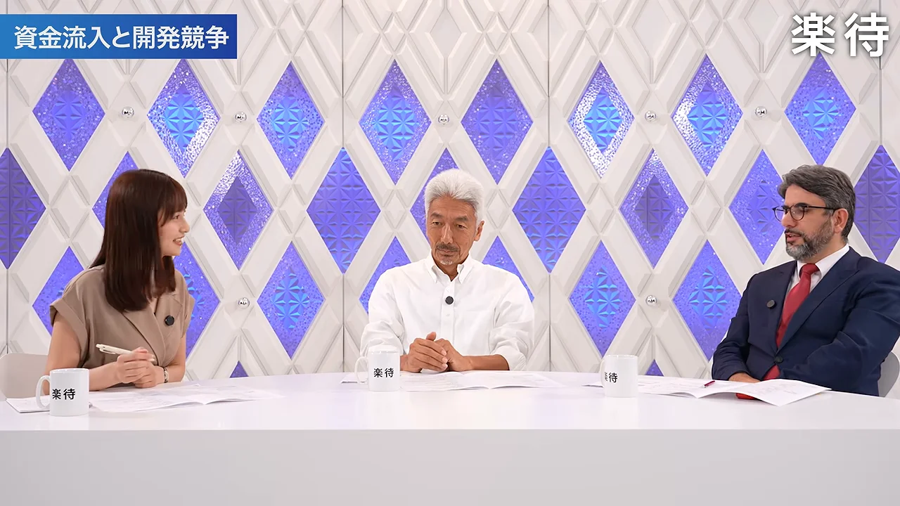 スタジオのテーブルで右側のスーツ姿の男性が話し、他の2人がその話を聞いています。