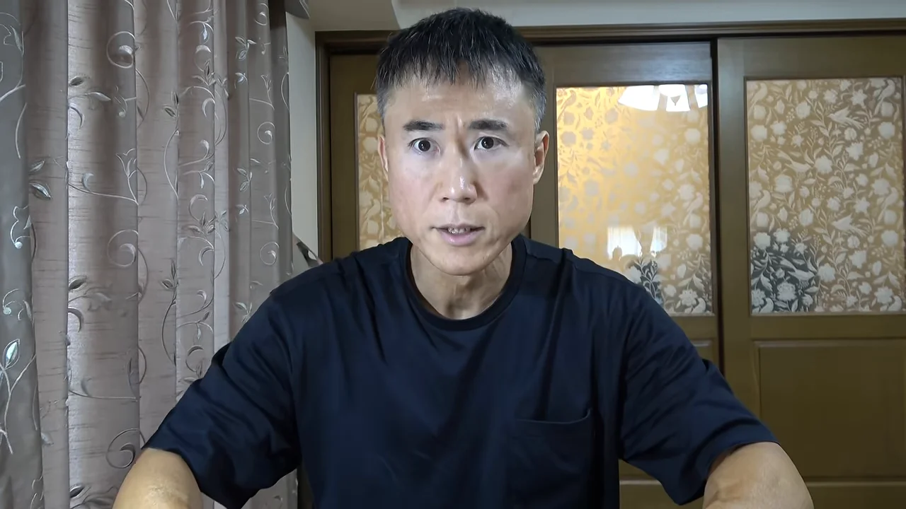 黒いTシャツを着た男性が、カーテンと引き戸を背景にカメラに向かって話しています。