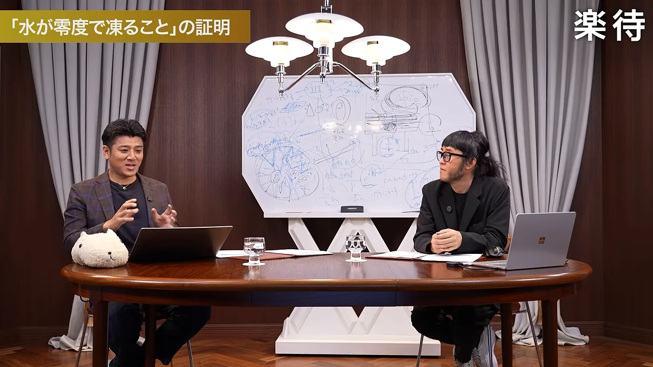 男性二人が机に座り、図や文字が描かれた大きなボードを背に話している。