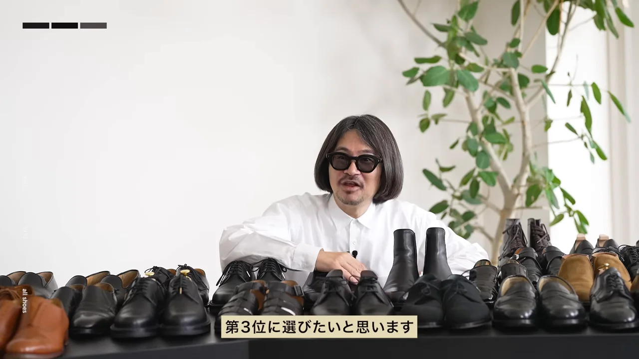テーブルいっぱいに並んだ革靴の前で身を乗り出しながら話す男性。