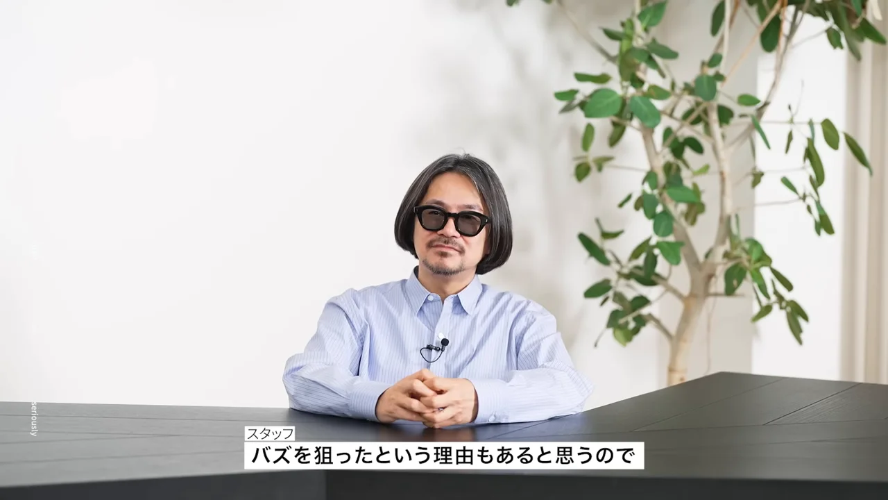 眼鏡をかけた男性が机の前で手を組んで座っている様子。
