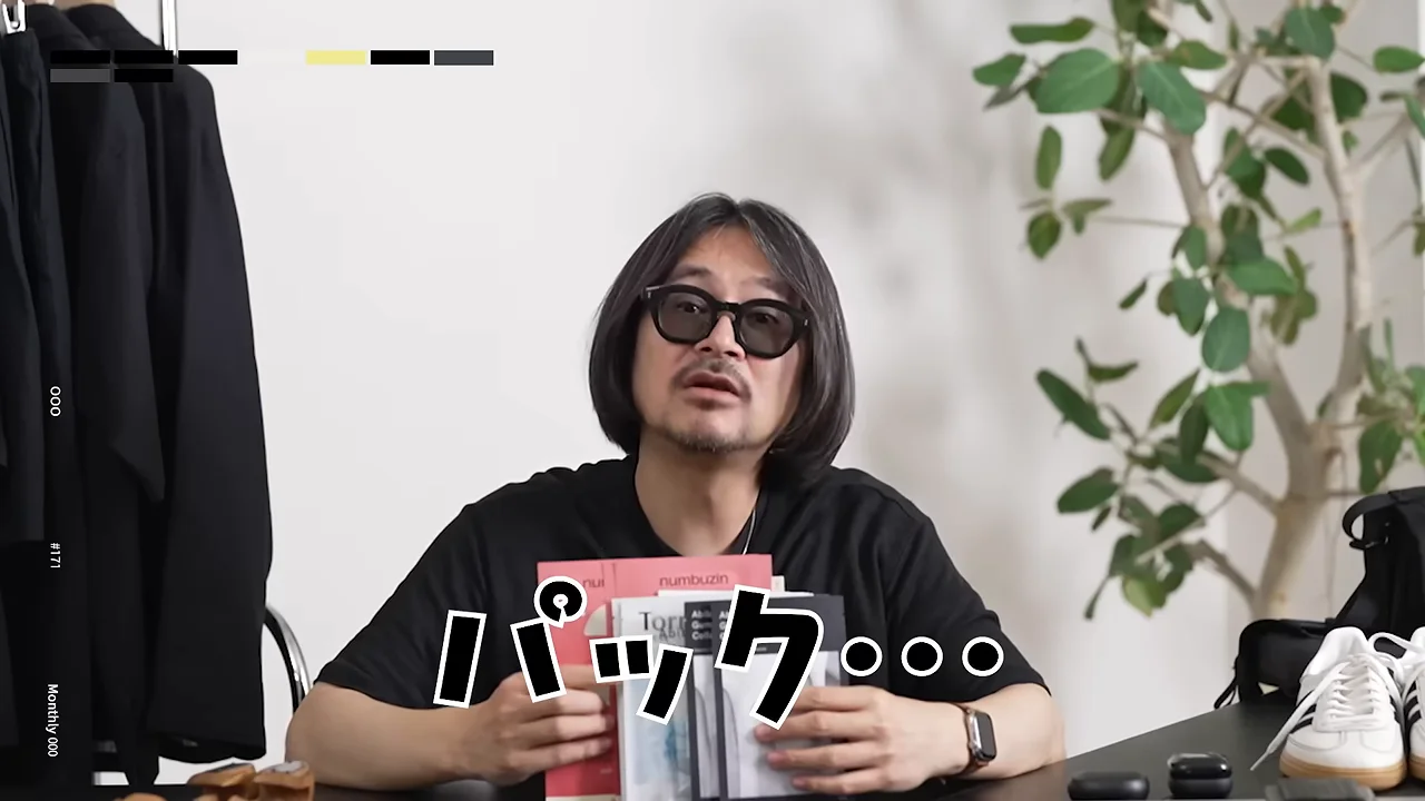 眼鏡をかけた男性がテーブルの前でフェイスパックのパッケージを手に持って話している様子。