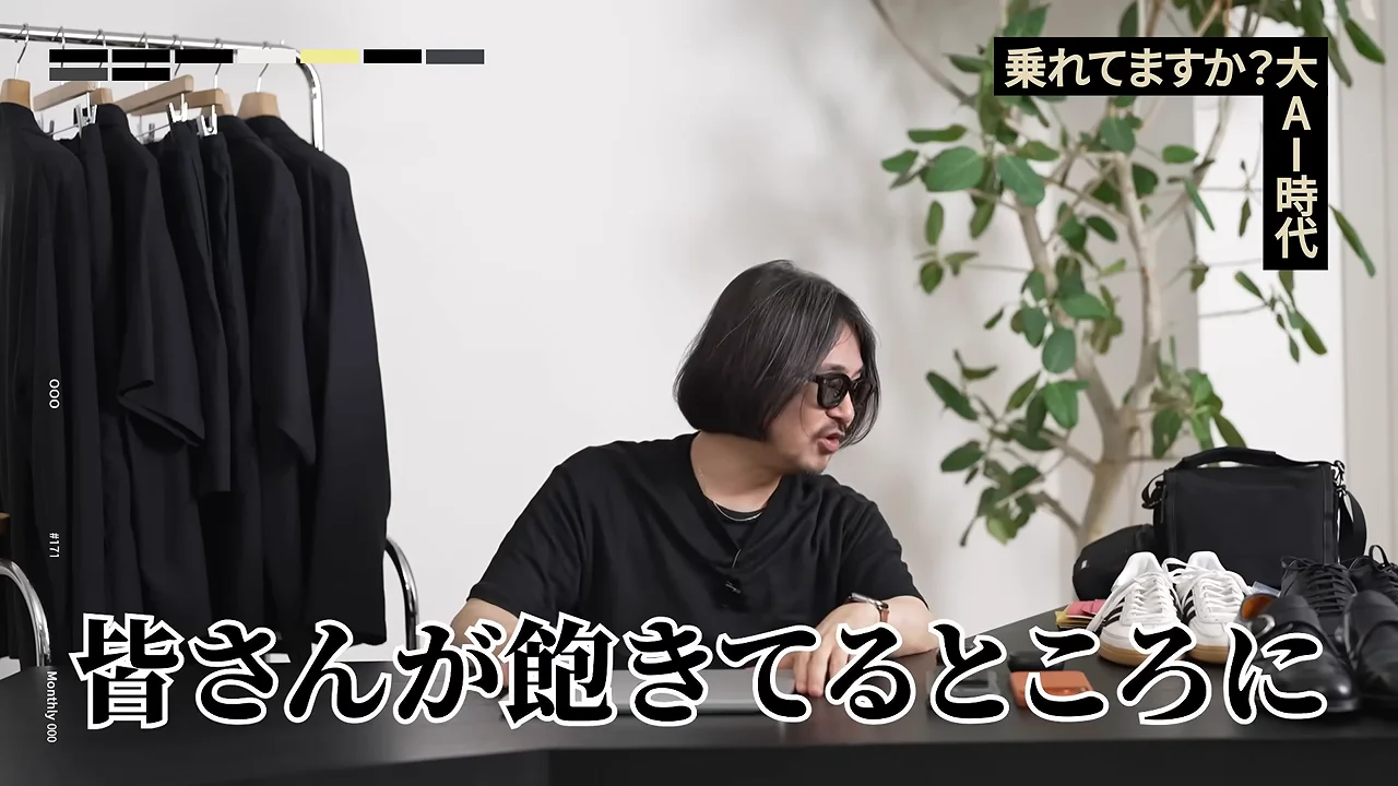 靴や小物が並ぶテーブルの前で、眼鏡をかけた男性が横を向いて話している様子。