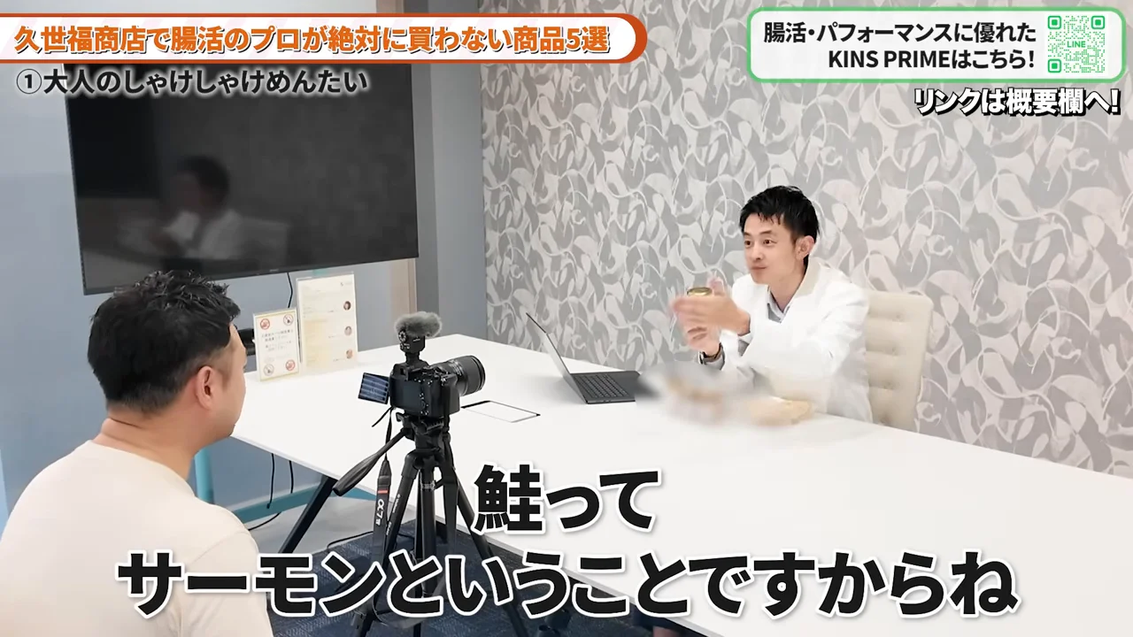 白い上着を着た男性がテーブルを挟んで向かいの人物と瓶を手に持ちながら話している様子。
