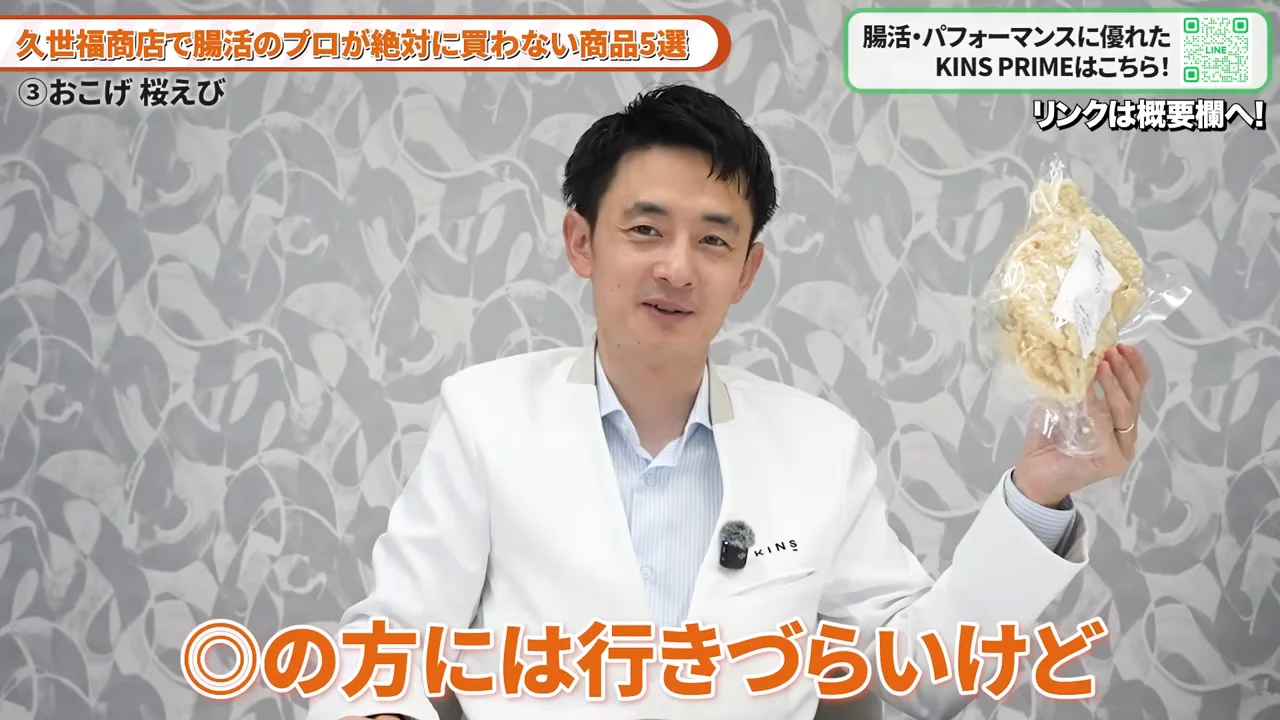 白衣姿の男性が透明な袋に入った菓子類を片手に持ち、カメラに向かって見せている様子。