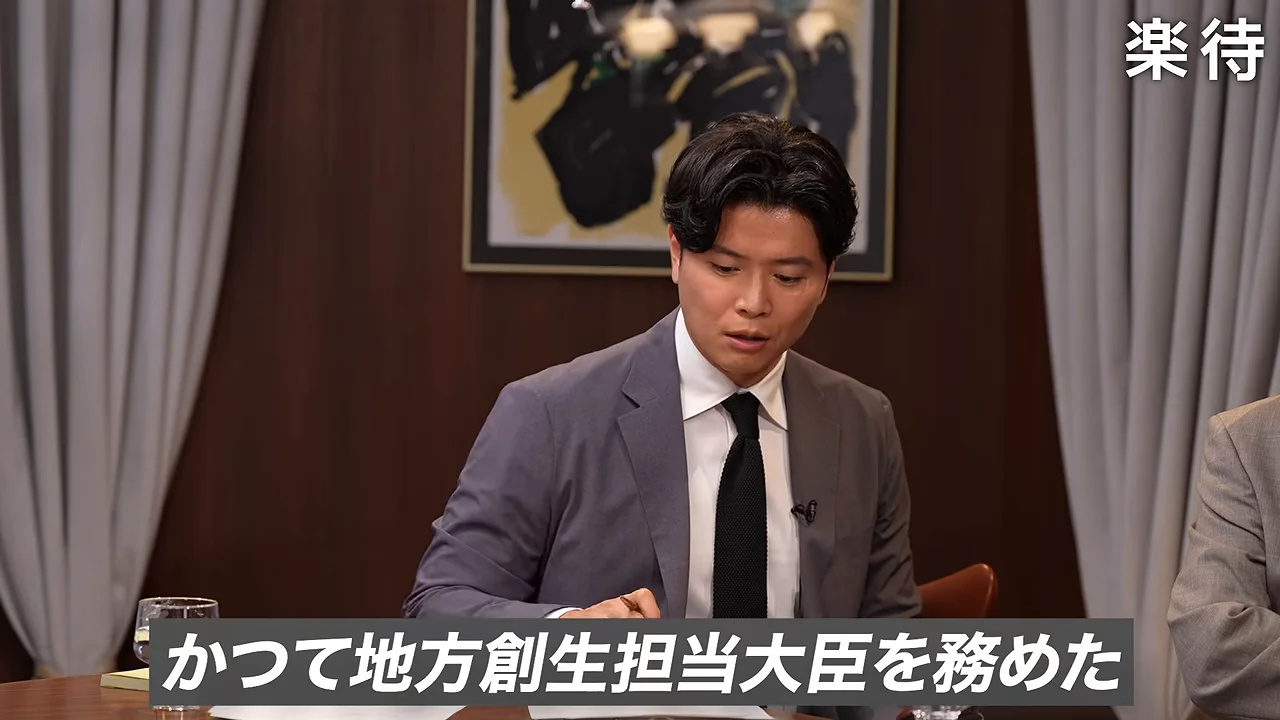 机に向かったスーツ姿の男性がペンを持ち手元を確認しながら語っています。